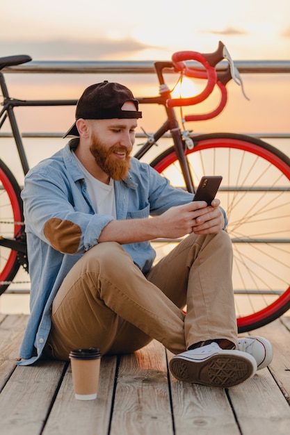 Bel homme barbu de style hipster portant chemise en jean et casquette tenant le smartphone avec vélo au lever du soleil du matin au bord de la mer, boire du café, voyageur de mode de vie actif sain