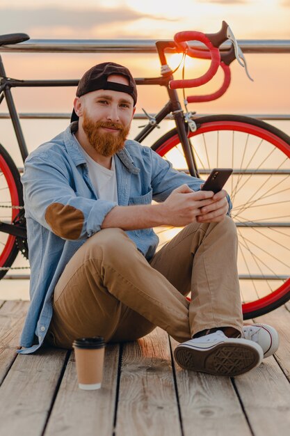 Bel homme barbu de style hipster portant chemise en jean et casquette tenant le smartphone avec vélo au lever du soleil du matin au bord de la mer, boire du café, voyageur de mode de vie actif sain