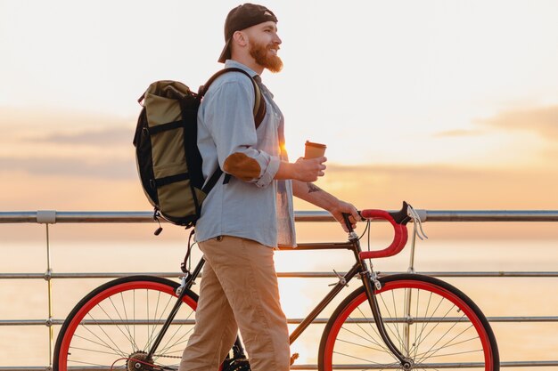 Bel homme barbu de style hipster avec sac à dos portant une chemise en jean et une casquette à vélo au lever du soleil du matin au bord de la mer, boire du café, routard de voyageur de mode de vie sain et actif