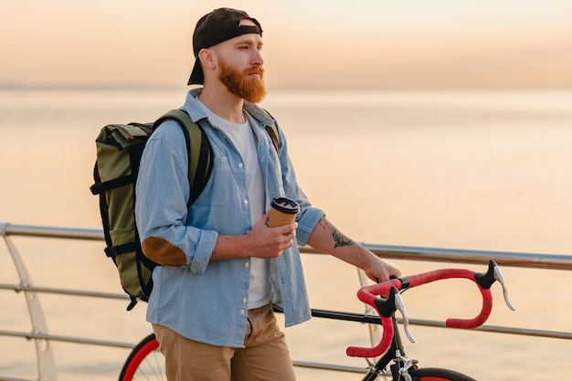 Bel homme barbu de style hipster avec sac à dos portant une chemise en jean et une casquette à vélo au lever du soleil du matin au bord de la mer, boire du café, routard de voyageur de mode de vie sain et actif