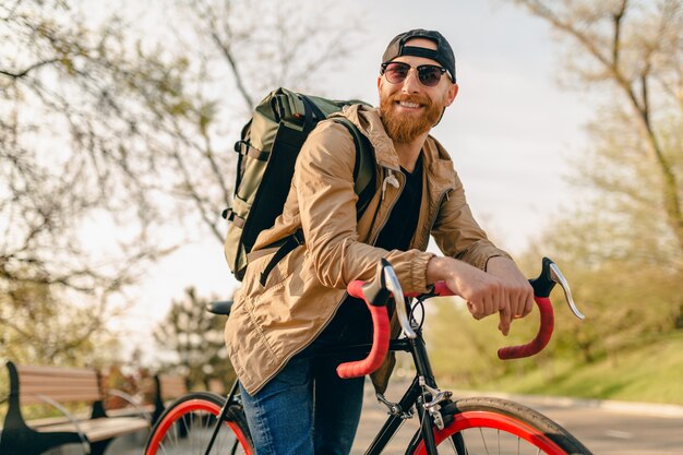 Bel homme barbu de style hipster en veste et lunettes de soleil à cheval seul avec sac à dos sur vélo routard voyageur de mode de vie actif sain