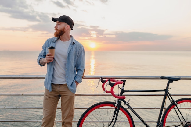 Bel Homme Barbu Voyageant à Vélo Au Lever Du Soleil Du Matin Au Bord De La Mer, Boire Du Café, Voyageur De Mode De Vie Actif Sain