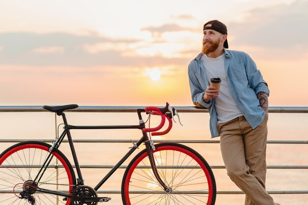 Bel homme barbu voyageant à vélo au lever du soleil du matin au bord de la mer, boire du café, voyageur de mode de vie actif sain