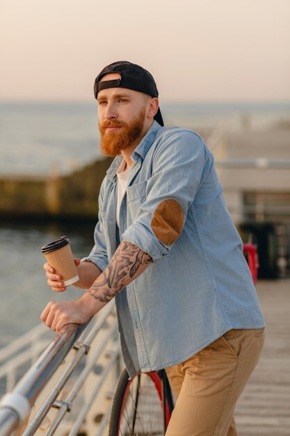 Bel homme barbu voyageant à vélo au lever du soleil du matin au bord de la mer, boire du café, voyageur de mode de vie actif sain