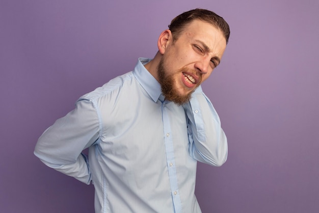 Bel homme blond douloureux tient le dos et le cou derrière isolé sur mur violet