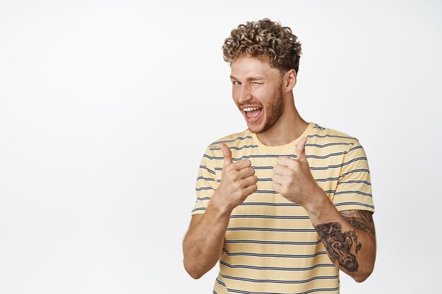 Bel homme blond fait un clin d'œil et montre les pouces vers le haut en signe d'approbation vous félicitant le gagnant debout sur fond blanc