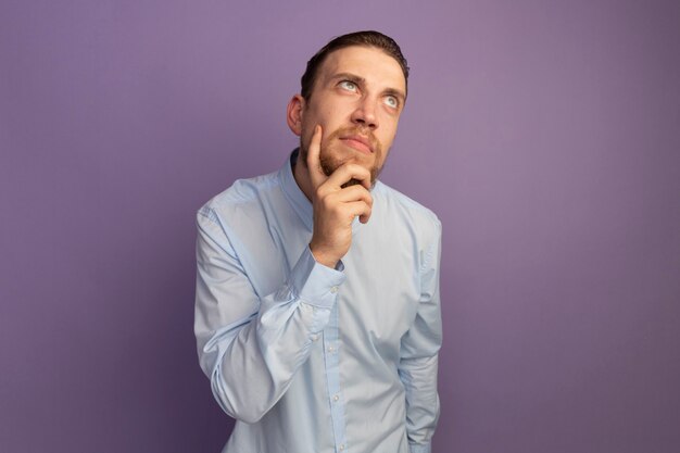 Bel homme blond réfléchi tient le menton et regarde le côté isolé sur le mur violet