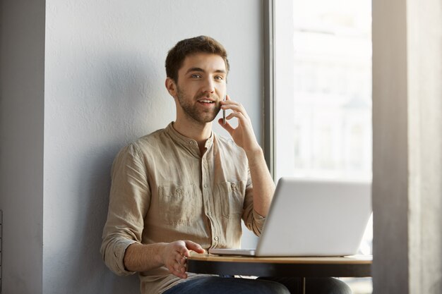 Bel homme caucasien aux cheveux noirs sourit, assis dans la cafétéria avec ordinateur portable, parler au téléphone et. Mode de vie, concept d'entreprise.