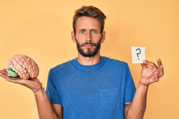 Bel homme caucasien avec barbe tenant le cerveau et le point d'interrogation sceptique et nerveux, fronçant les sourcils contrarié à cause du problème. personne négative.