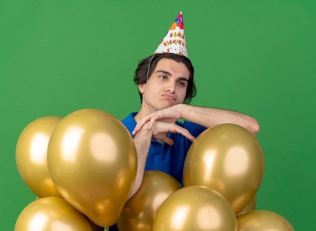 Un bel homme caucasien bouleversé portant une casquette d'anniversaire se tient avec des ballons à l'hélium