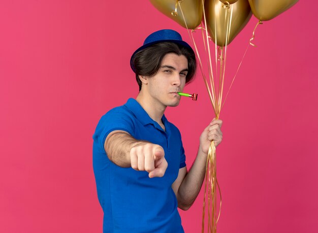 Un bel homme caucasien confiant portant un chapeau de fête bleu tient des ballons à l'hélium soufflant un sifflet de fête et pointant vers la caméra