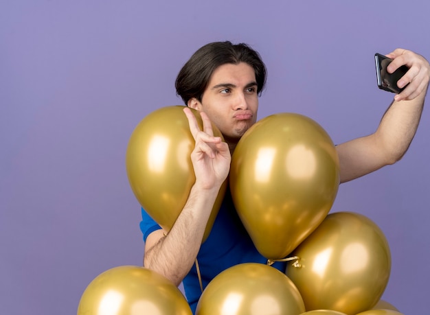 Un bel homme caucasien confiant se tient avec des ballons à l'hélium gesticulant le signe de la main de la victoire et regardant le téléphone prenant un selfie