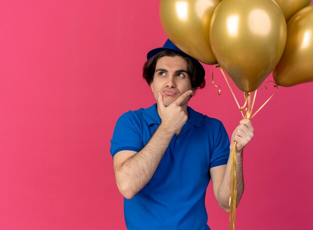 Un bel homme caucasien confus portant un chapeau de fête bleu tient des ballons à l'hélium met la main sur le menton