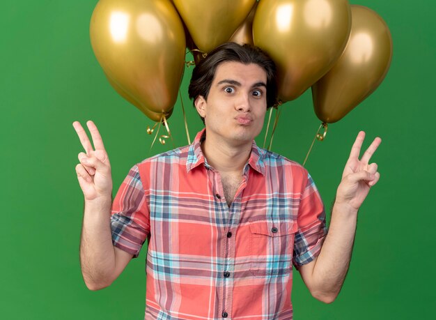 Un bel homme caucasien impressionné portant une casquette d'anniversaire se tient devant des ballons à l'hélium gesticulant le signe de la main de la victoire à deux mains