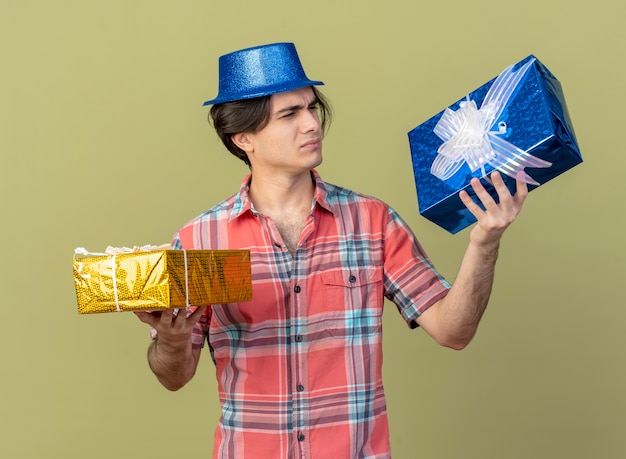 Un bel homme caucasien mécontent portant un chapeau de fête bleu tient et regarde les coffrets cadeaux