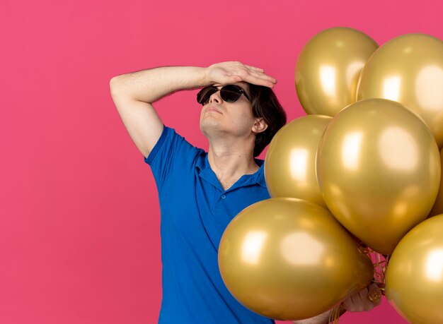 Un bel homme caucasien offensé à lunettes de soleil tient des ballons à l'hélium et met la main sur le front