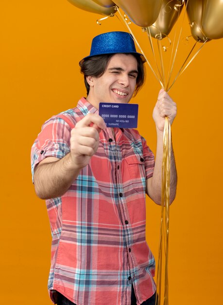 Un bel homme caucasien souriant et confiant portant un chapeau de fête bleu tient des ballons à l'hélium et une carte de crédit