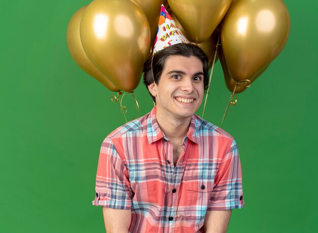 Un bel homme caucasien souriant portant une casquette d'anniversaire se tient devant des ballons à l'hélium regardant la caméra