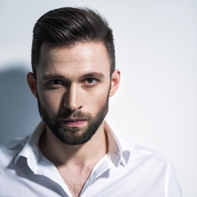 Bel homme en chemise blanche, posant. Mec attrayant avec une coiffure de mode. Homme confiant avec une barbe courte. Garçon adulte aux cheveux bruns. Portrait en gros plan.