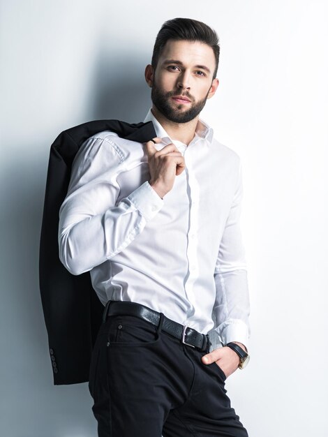 Bel homme en chemise blanche tient le costume noir - posant sur le mur. Mec attrayant avec une coiffure de mode. Homme confiant avec une barbe courte. Garçon adulte aux cheveux bruns. Portrait complet.