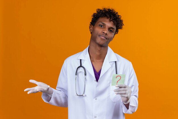 Un bel homme confus à la peau sombre avec des cheveux bouclés portant blouse blanche avec stéthoscope montrant une carte en papier avec le point d'interrogation