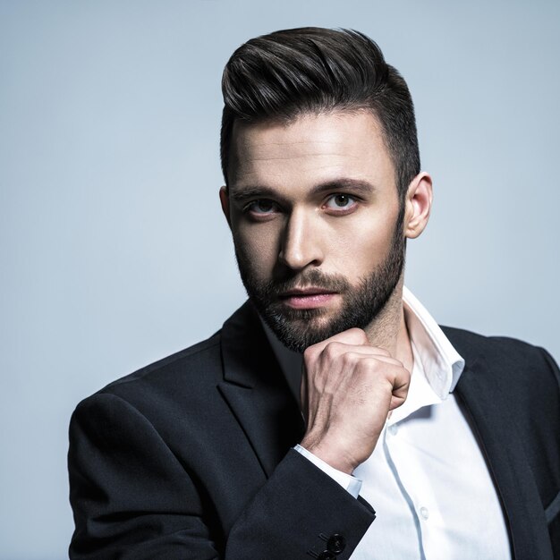 Bel homme en costume noir avec chemise blanche - posant Beau mec avec une coiffure de mode. Homme confiant avec une barbe courte. Garçon adulte aux cheveux bruns.