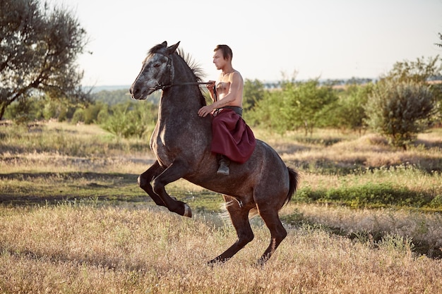 Photo gratuite bel homme cow-boy à cheval sur un cheval - fond de ciel et d'arbres. un homme en pantalon large rouge sans chemise. spectacle