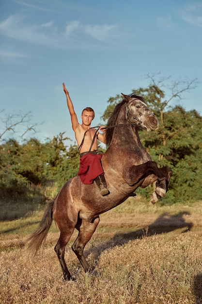 Bel homme cow-boy à cheval sur un cheval - fond de ciel et d'arbres. Un homme en pantalon large rouge sans chemise. Spectacle