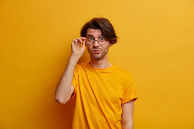 bel homme curieux regarde attentivement à travers des lunettes, a un regard attentif, vêtu de vêtements décontractés, a un regard scrupuleux, isolé sur un mur jaune, reçoit une suggestion intéressante