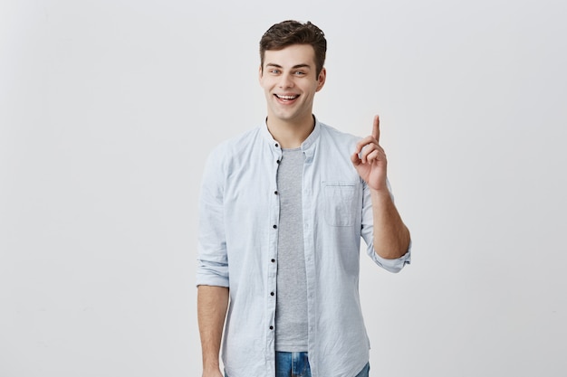 Bel homme élégant aux yeux bleus attrayants, souriant à la caméra avec une expression heureuse pointant avec les index vers l'espace vide au-dessus de la tête pour votre contenu promotionnel. Concept publicitaire.