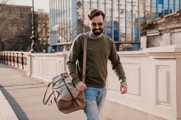 Bel homme élégant hipster marchant dans la rue de la ville avec sac en cuir portant sweatshot et lunettes de soleil, tendance de style urbain, journée ensoleillée, souriant voyageur heureux