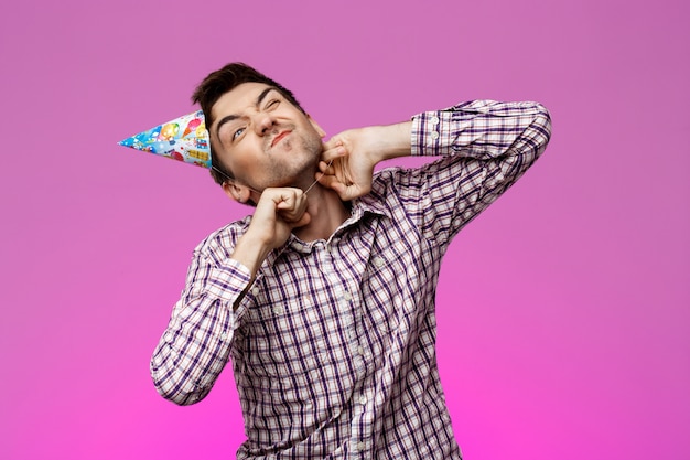 Bel homme essayant de décoller le chapeau d'anniversaire sur le mur violet.