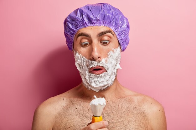 Un bel homme étonné regarde la brosse, le gel à raser Appiles, veut avoir une peau lisse, a des procédures de beauté à la maison, porte un chapeau spécial