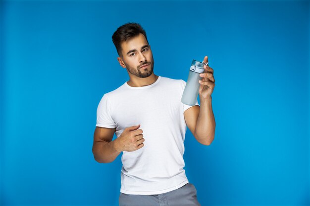 Bel homme européen en t-shirt blanc sur fond bleu tient une bouteille de sport dans une main