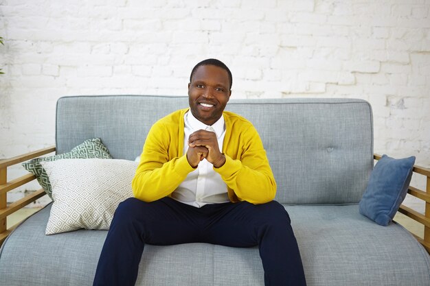 Bel homme gai à la peau sombre portant des jeans et un cardigan jaune, assis sur un canapé confortable dans le salon, serrant les mains et souriant largement, montrant ses dents blanches parfaites