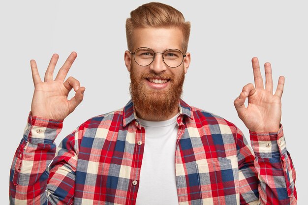 Bel homme heureux satisfait aime le plan de quelqu'un, montre un signe correct, des gestes en studio, a une barbe épaisse rouge et une coupe de cheveux