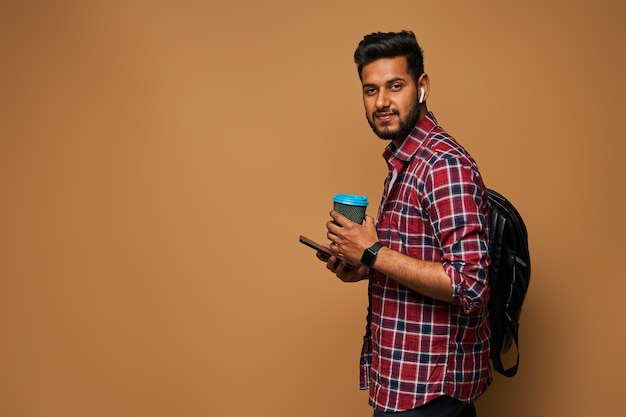 Bel homme hindou regardant vers l'avant avec du café à emporter et un sac à dos sur un mur pastel