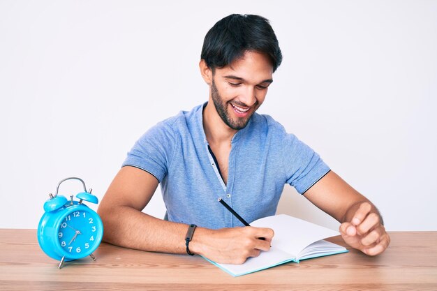 Bel homme hispanique assis sur la table étudiant pour l'université à la recherche positive et heureuse debout et souriant avec un sourire confiant montrant les dents