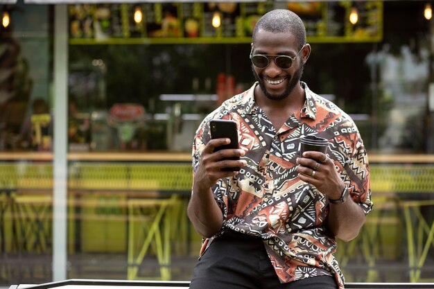 Bel homme avec des lunettes de soleil à l'aide d'un smartphone à l'extérieur tout en prenant un café