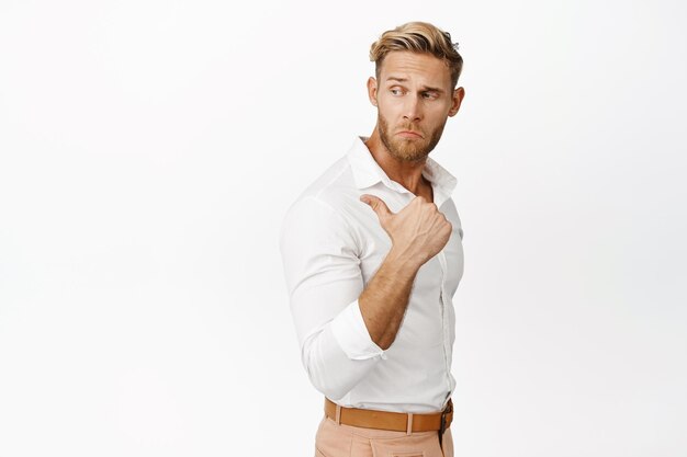 Bel homme macho en chemise blanche pointant et regardant derrière son épaule montrant le produit promotionnel de vente sur fond de studio espace copie vide