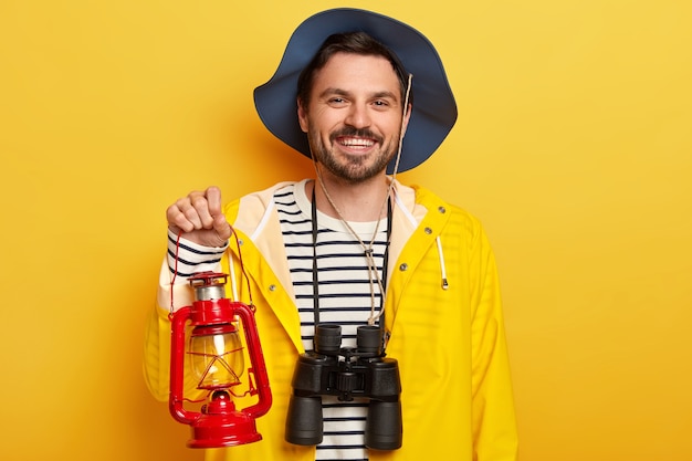 Bel homme mal rasé porte une lampe à pétrole, des jumelles, prêt pour une expédition ou un voyage, porte un chapeau et un imperméable, isolé sur un mur jaune