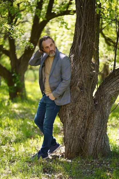 Un bel homme mature aux oiseaux d'âge mûr en position décontractée sous l'arbre sur l'herbe regardant derrière l'arbre avec les mains dans les poches Le pigiste passe du temps à travailler à l'extérieur