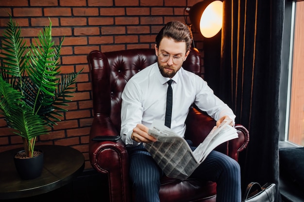 Bel homme millionnaire élégant en costume bleu à la maison assis sur un canapé et lisant le journal.