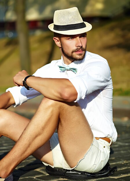 Bel homme modèle hipster dans des vêtements d'été élégants assis dans un chapeau avec sac
