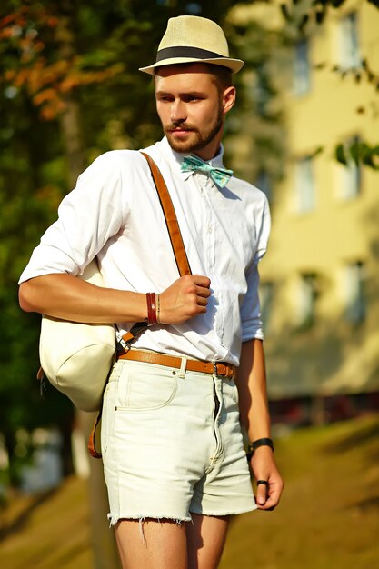 Bel homme modèle hipster dans des vêtements d'été élégants posant en chapeau avec sac