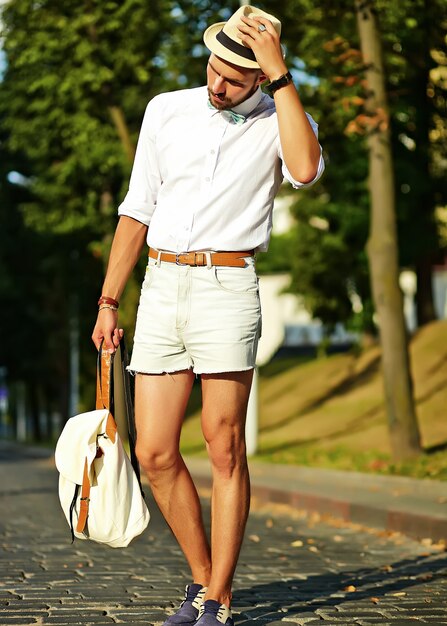 Bel homme modèle hipster dans des vêtements d'été élégants posant en chapeau avec sac