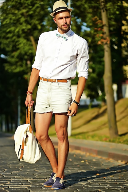 Bel homme modèle hipster dans des vêtements d'été élégants posant en chapeau avec sac