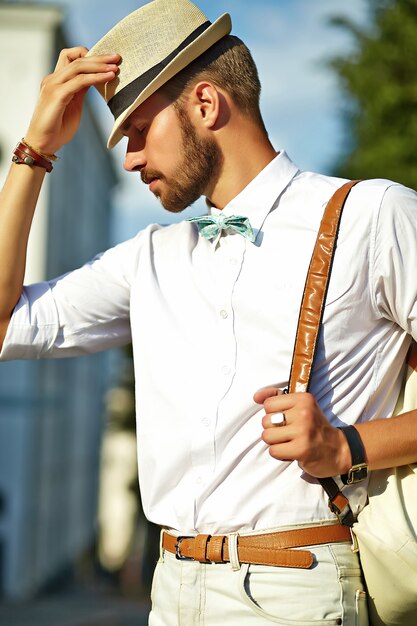 Bel homme modèle hipster dans des vêtements d'été élégants posant en chapeau avec sac