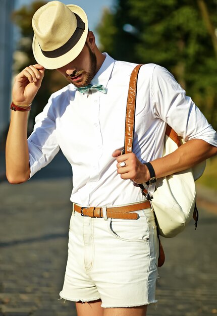 Bel homme modèle hipster dans des vêtements d'été élégants posant en chapeau avec sac