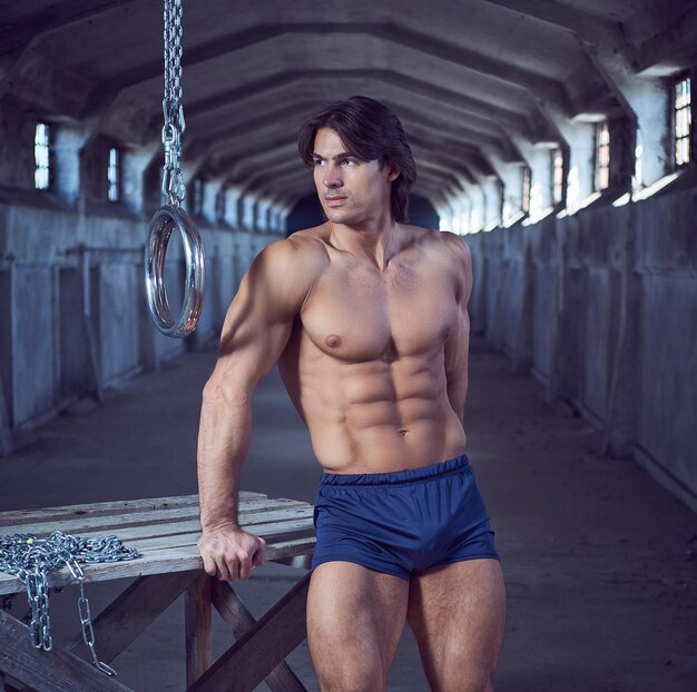 Bel homme musclé avec un corps sportif debout tout en s'appuyant sur une table en bois près d'anneaux en acier dans le tunnel sombre.